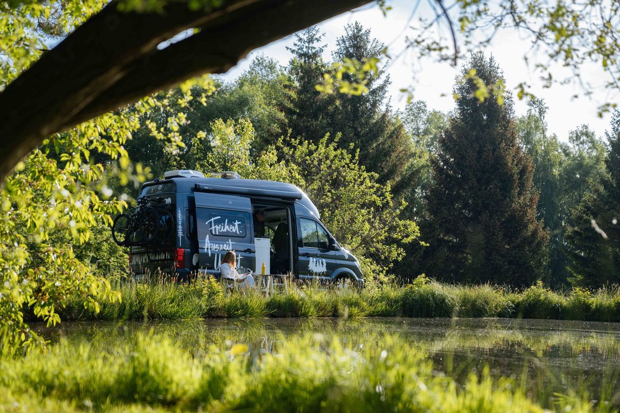 Camper Van steht auf einem Camping Stellplatz direkt am Wasser in Alleinlage umgeben von grünen Wiesen, Wald, einem See und einem Wandergebiet, inseriert auf hinterland.camp