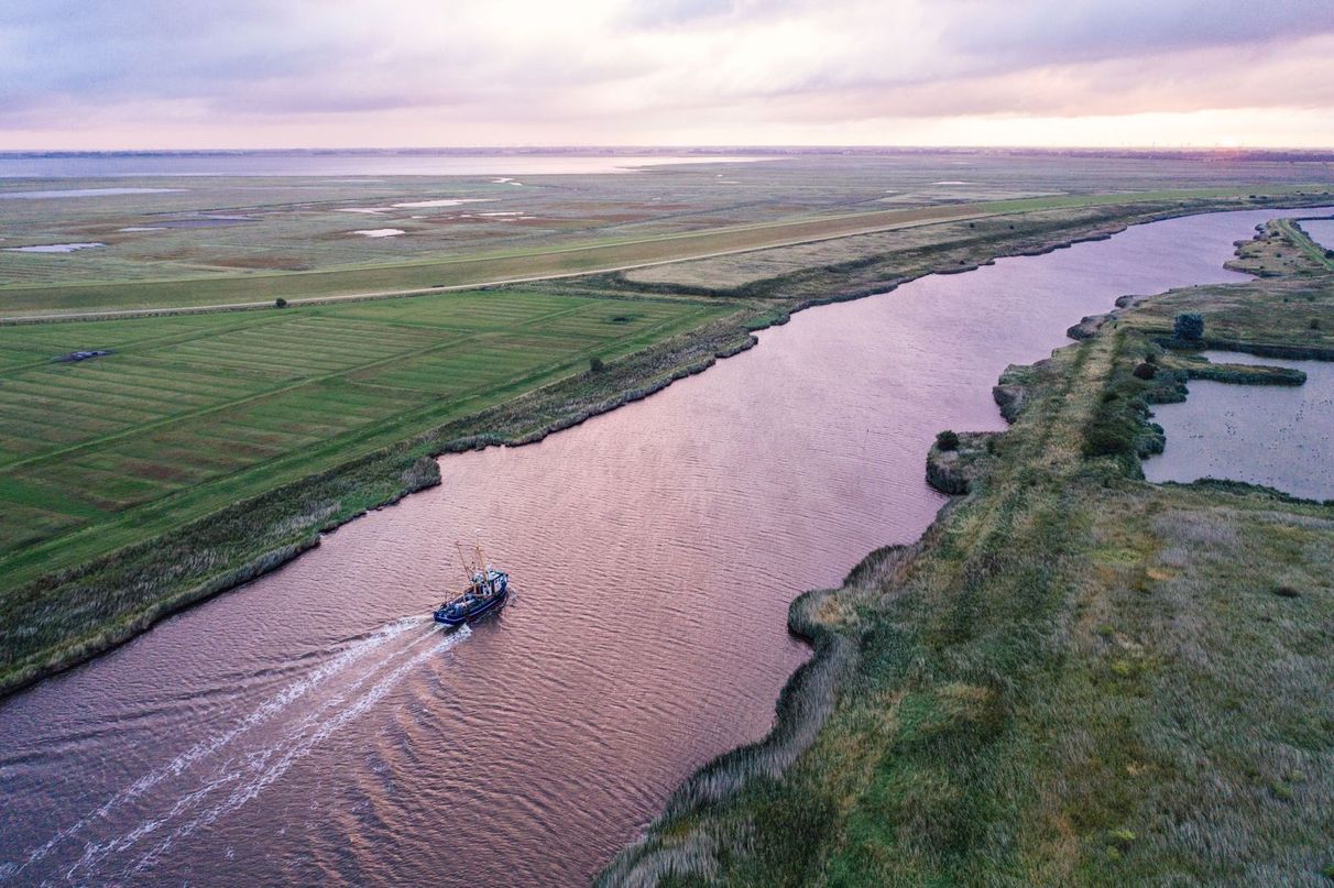 Sonnenuntergang, Drohnen-Aufnahme eines Flusses, auf dem ein Boot fährt, von Wiesen umgeben