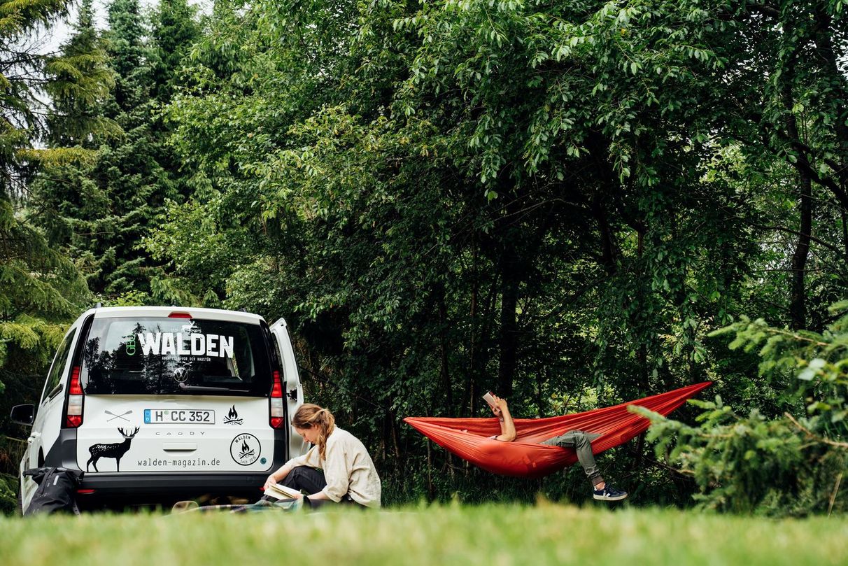 Eine Person liegt in der Hängematte und liest dabei ein Buch, eine weitere Person sitzt auf dem Rasen, beide befinden sich vor dem VW-Camper von Walden auf einem Platz von hinterland.camp