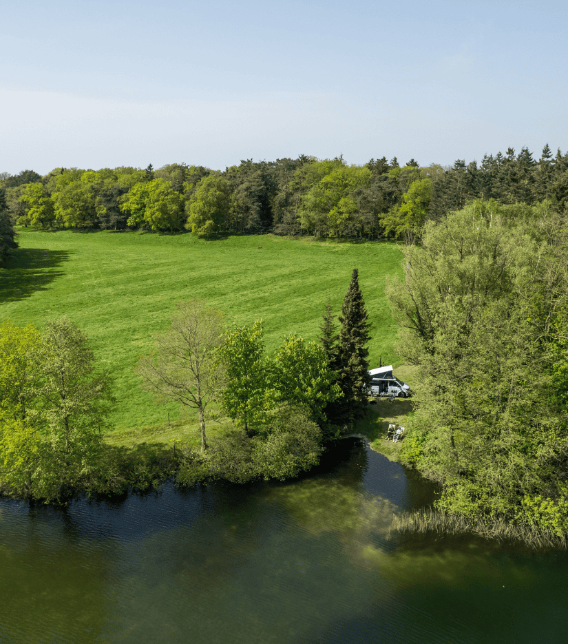 VW Van right in front of a lake between forest and meadows.