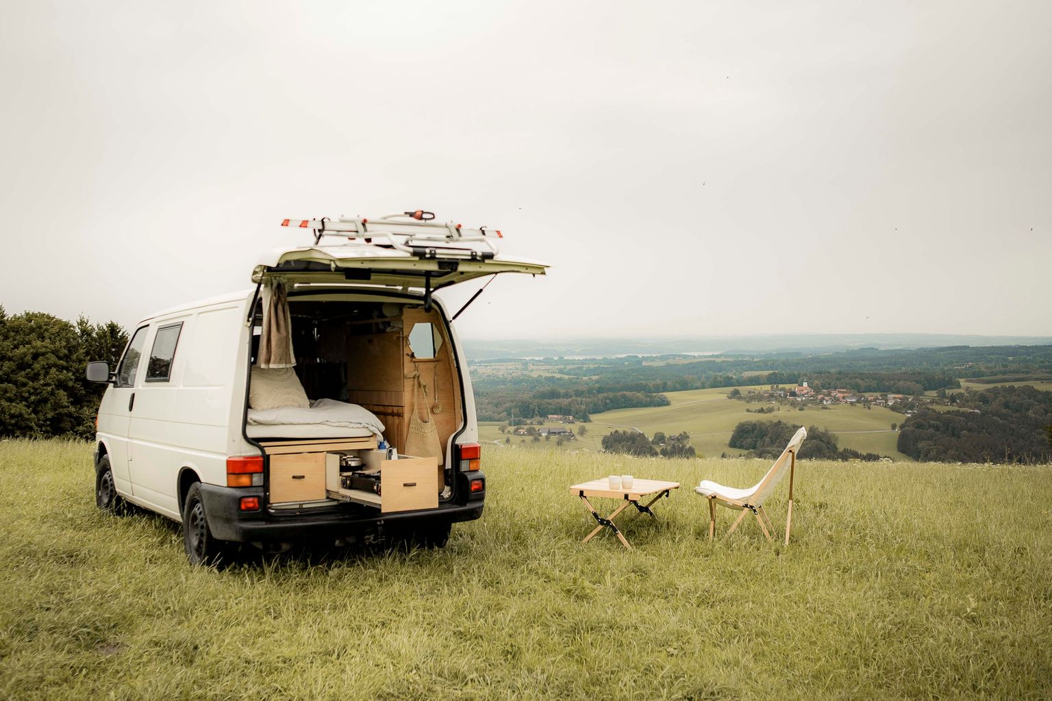 Camper mit offener Heckklappe steht auf einer Wiese mit wunderschönem Blick über die Berge Bayerns. Es stehen Campingstühle draußen.