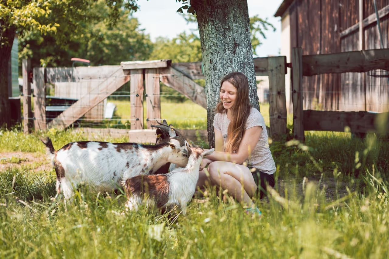 Eine Frau hockt auf einer Weide vor einem Gatter umgeben von einer Zwergziegenherde. Die Wiese gehört zu dem Abenteuer-Campingplatz, den man auf hinterland.camp buchen kann.