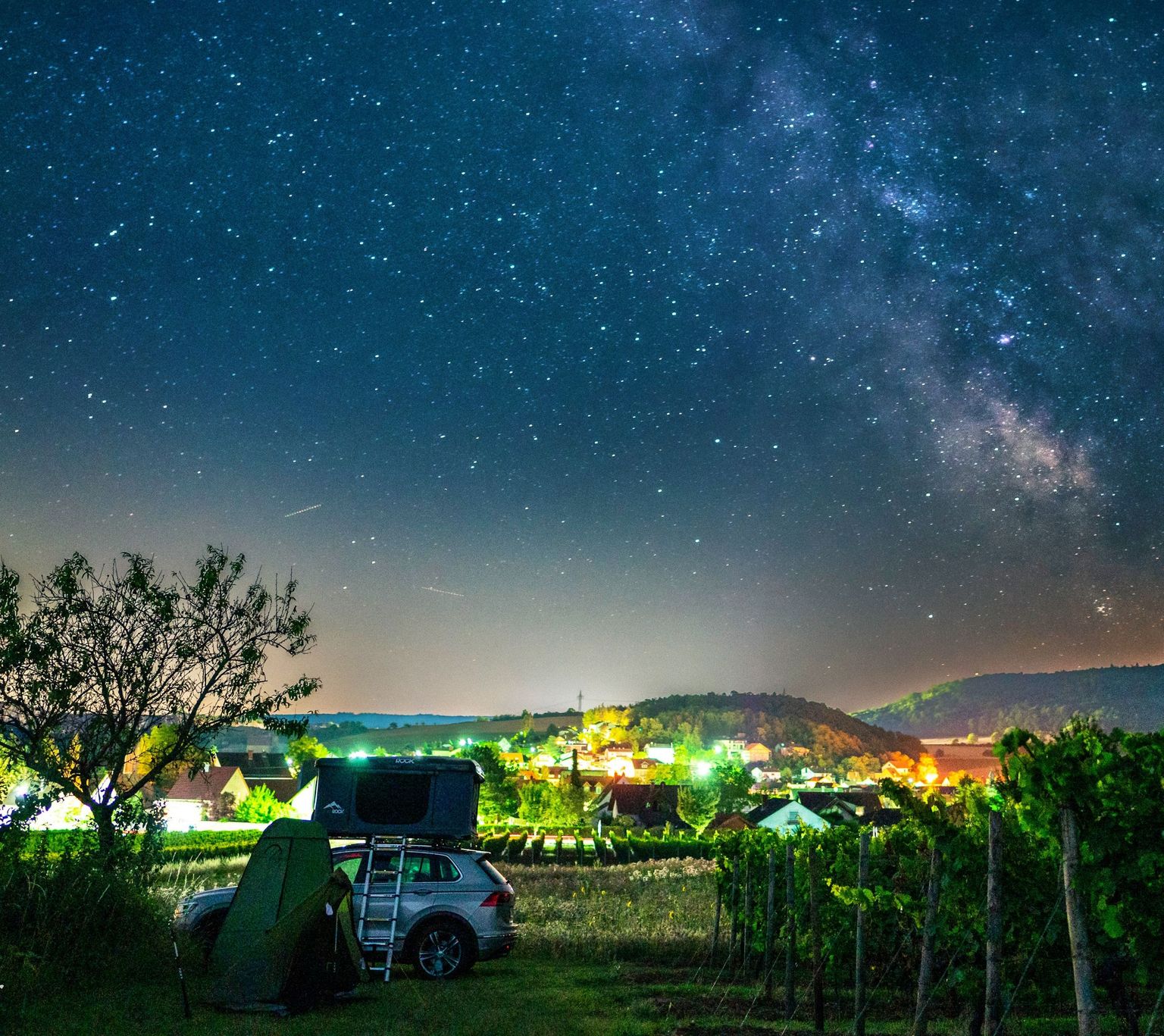 Ein Camper steht bei Nacht auf einem Grünstreifen direkt neben den Weinreben. Man hat einen wunderschönen Panoramablick über die leuchtende Weinregion.