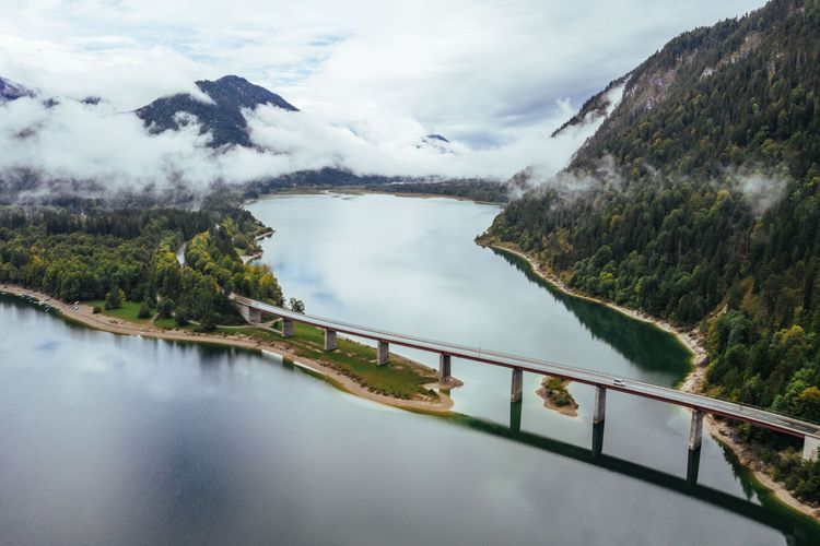 Eine Fluss in den Alpen, der die Berge durchquert. Eine Verbindung der Berge stellt eine befahrbare Brücke dar, über die ein Camper in den Wald fährt