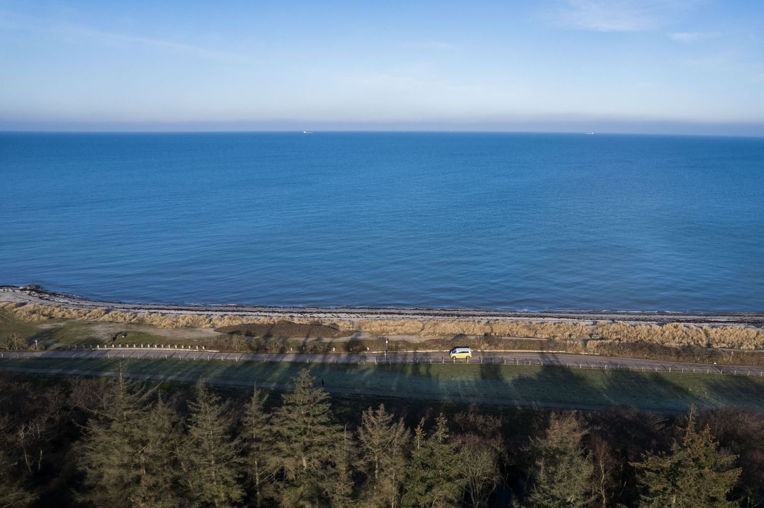 Drohnen-Aufnahme eines Campers, der eine Straße entlang der Ostsee fährt, direkt am Meer der Ostsee, umgeben von einem Wald