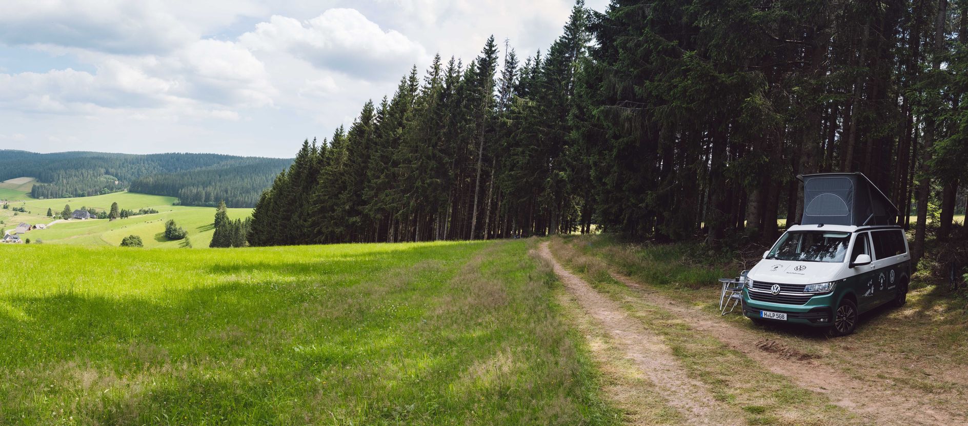 Naturcampingplatz in der Eifel, Camper steht vor einem Kiefernwald an einer Wiese auf einem Stellplatz von hinterland.camp mit Panoramablick auf ein Tal in der Eifel