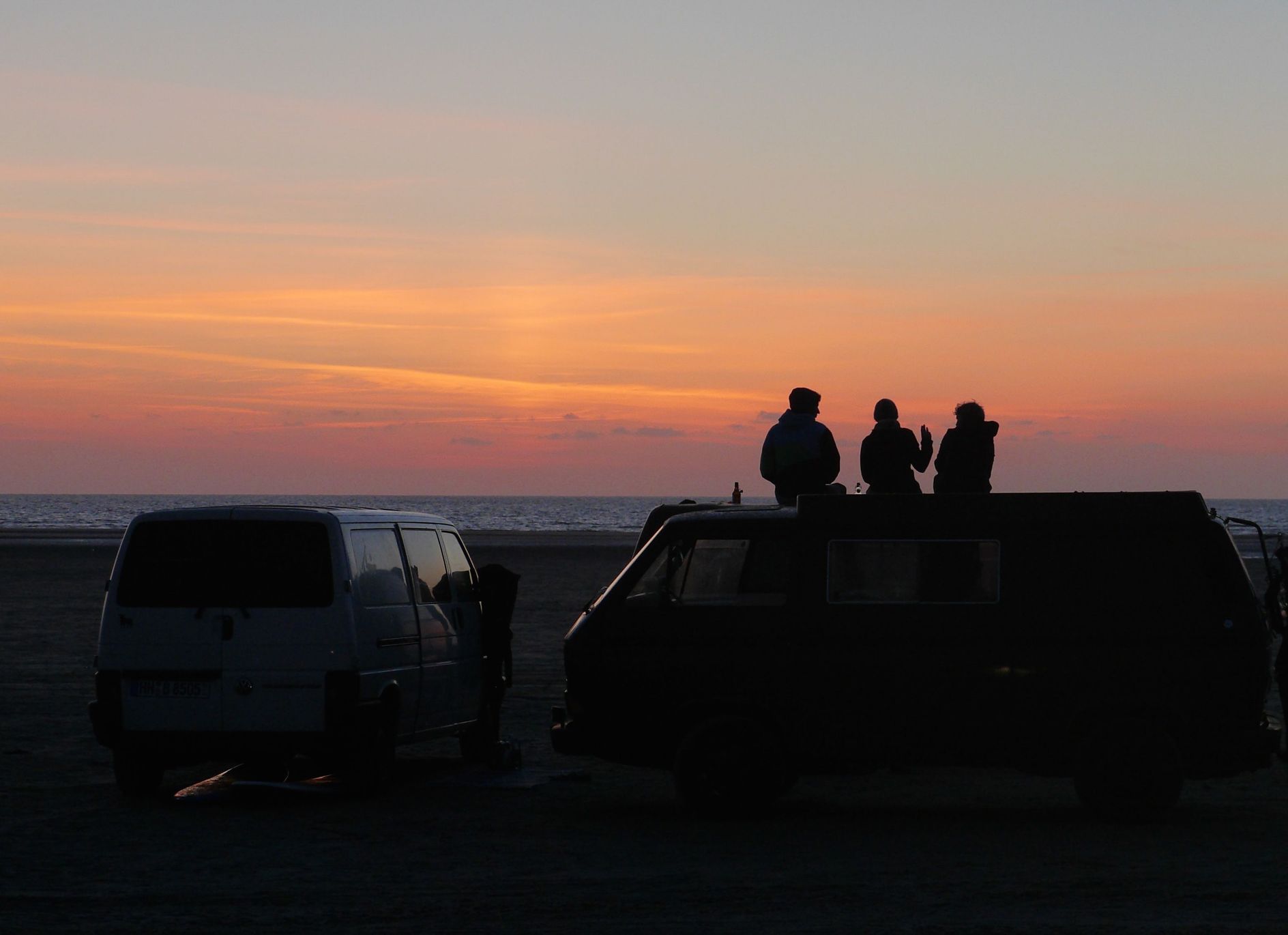 3 Bulli stehen direkt am Strand der Ostsee in Schleswig Holstein. Drei Menschen sitzen auf der Dachterasse eines T3 Campers direkt am Strand in Alleinlage