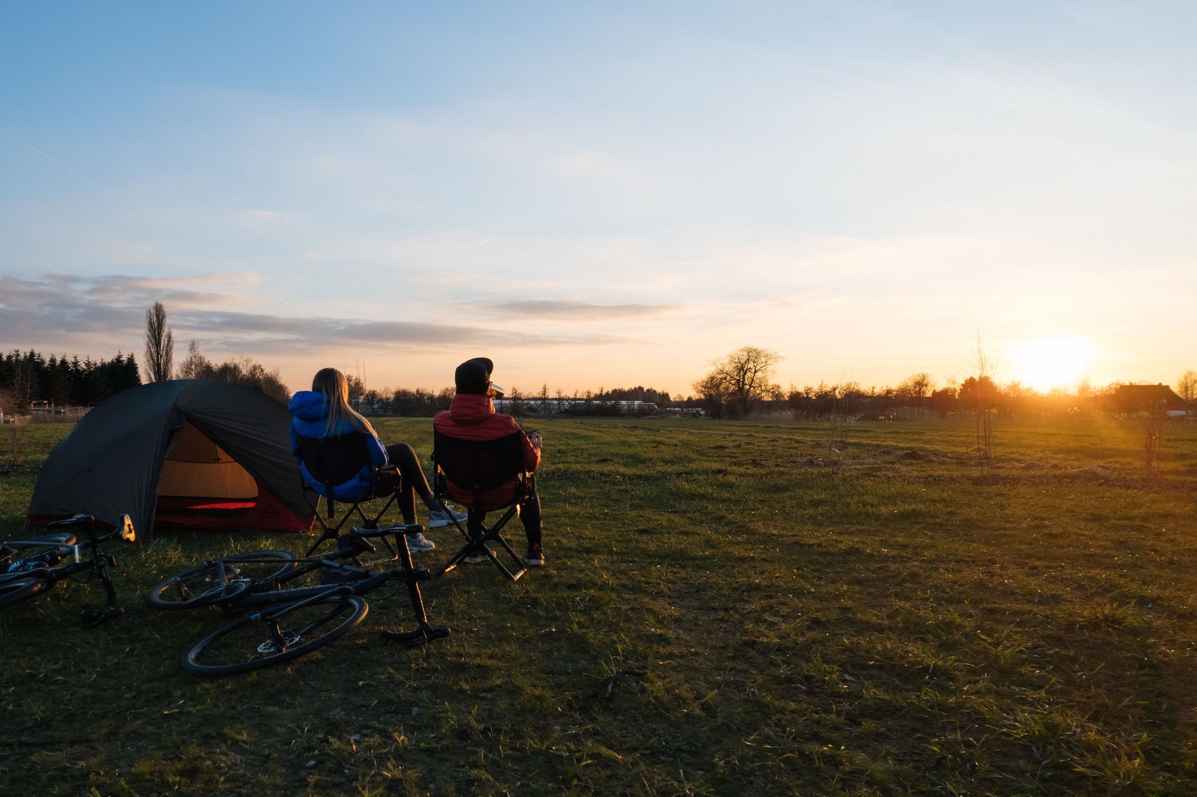 Eine Frau und ein Mann sitzen auf Campingstühlen neben ihrem Zelt bei Sonnenuntergang. Sie zelten auf einem naturnahen Zeltplatz in der Nähe von Hamburg.