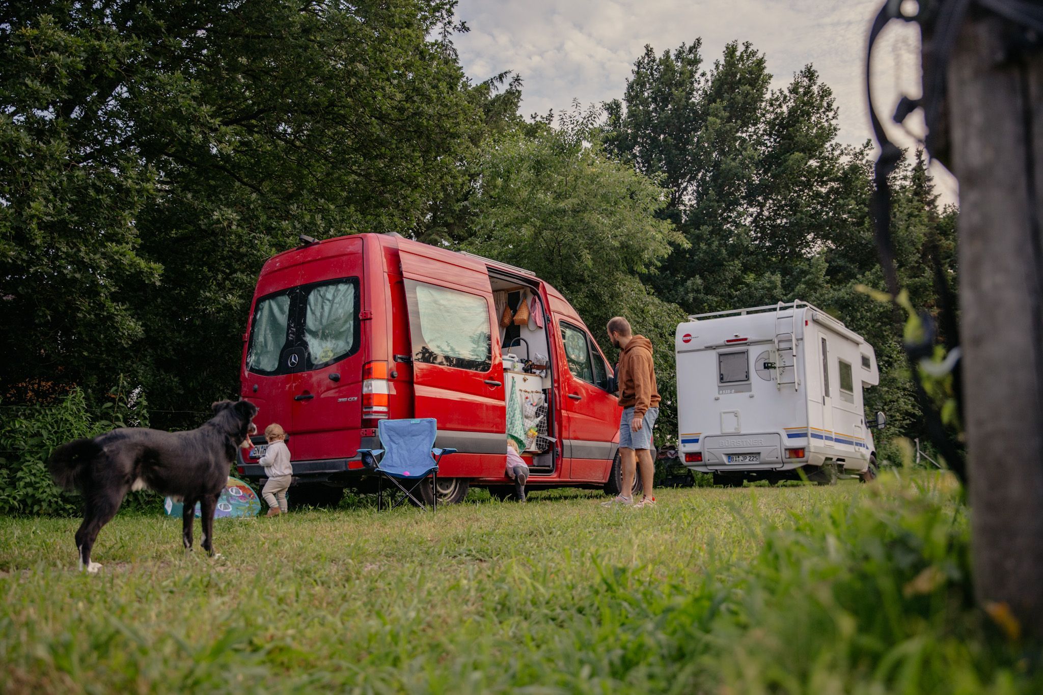 Zwei Camper stehen vor einer Baumreihe auf einer Wiese auf einem Camping Stellplatz von hinterland.camp in der Sächsischen Schweiz. Davor sieht man Kinder und einen Hund die auf dem Rasen spielen und Eltern, die am Camper stehen.