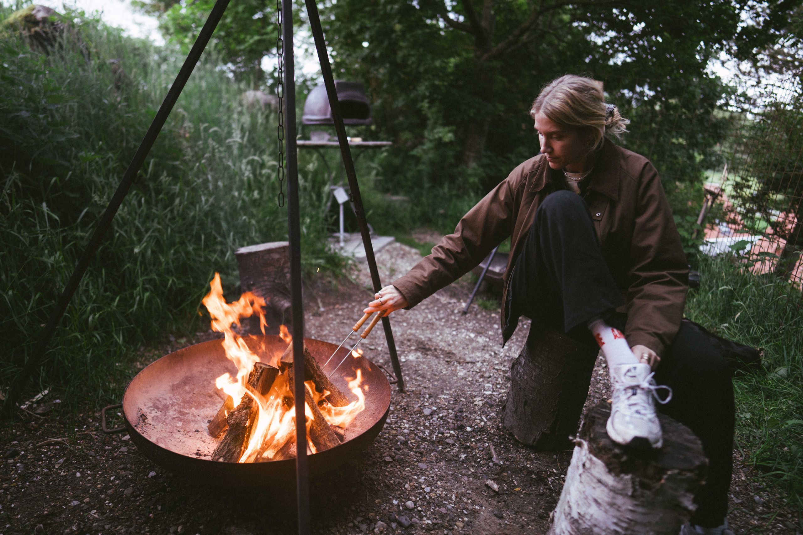 Eine Frau sitzt vor einem Schwenkgrill, welcher zu einer naturnahen Hütte im Wald gehört, die man über hinterland.camp buchen kann.