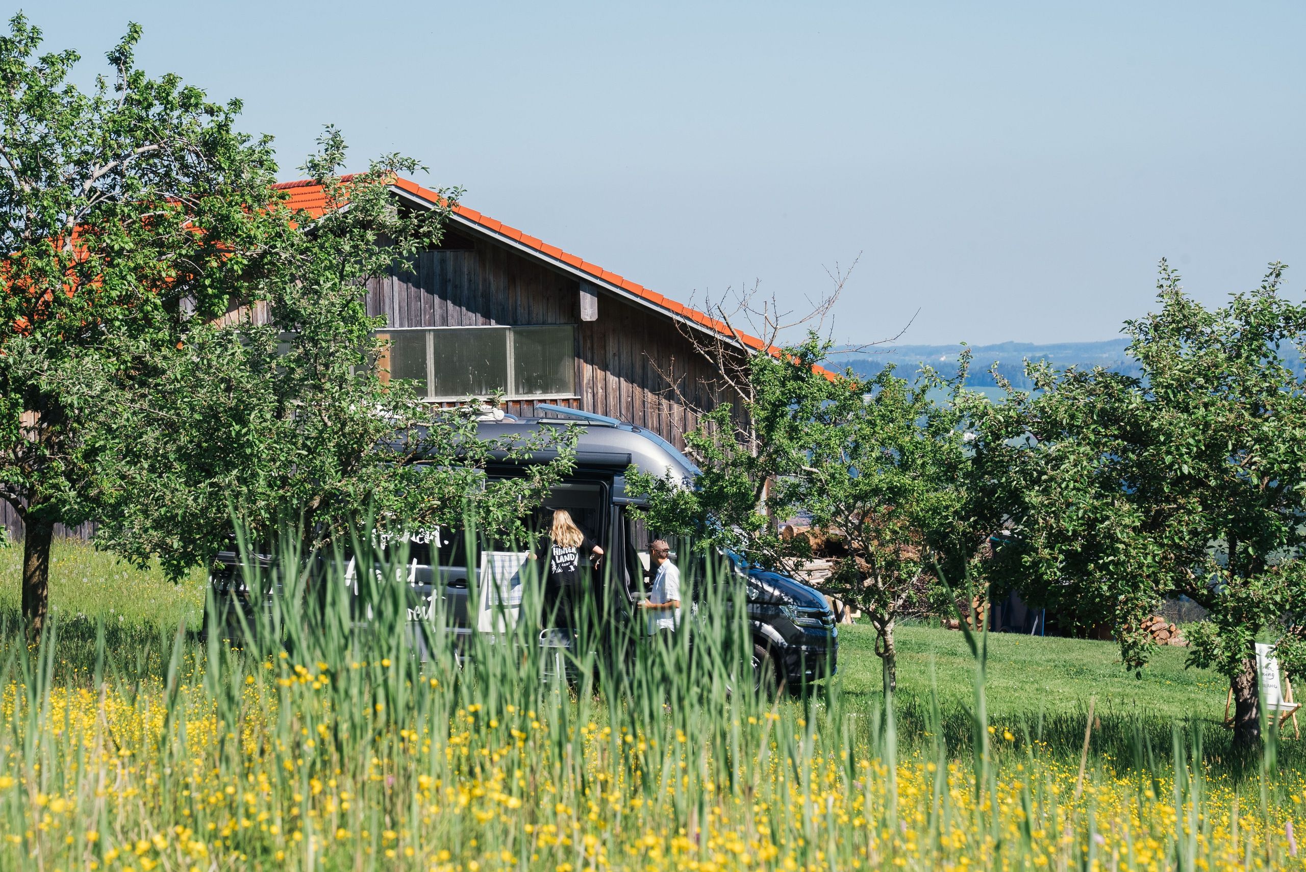 Ein Grand California Camper steht auf einer Blumenwiese auf einem Bauernhof. Im Hintergrund sieht man eine Scheune des Bauernhofs.