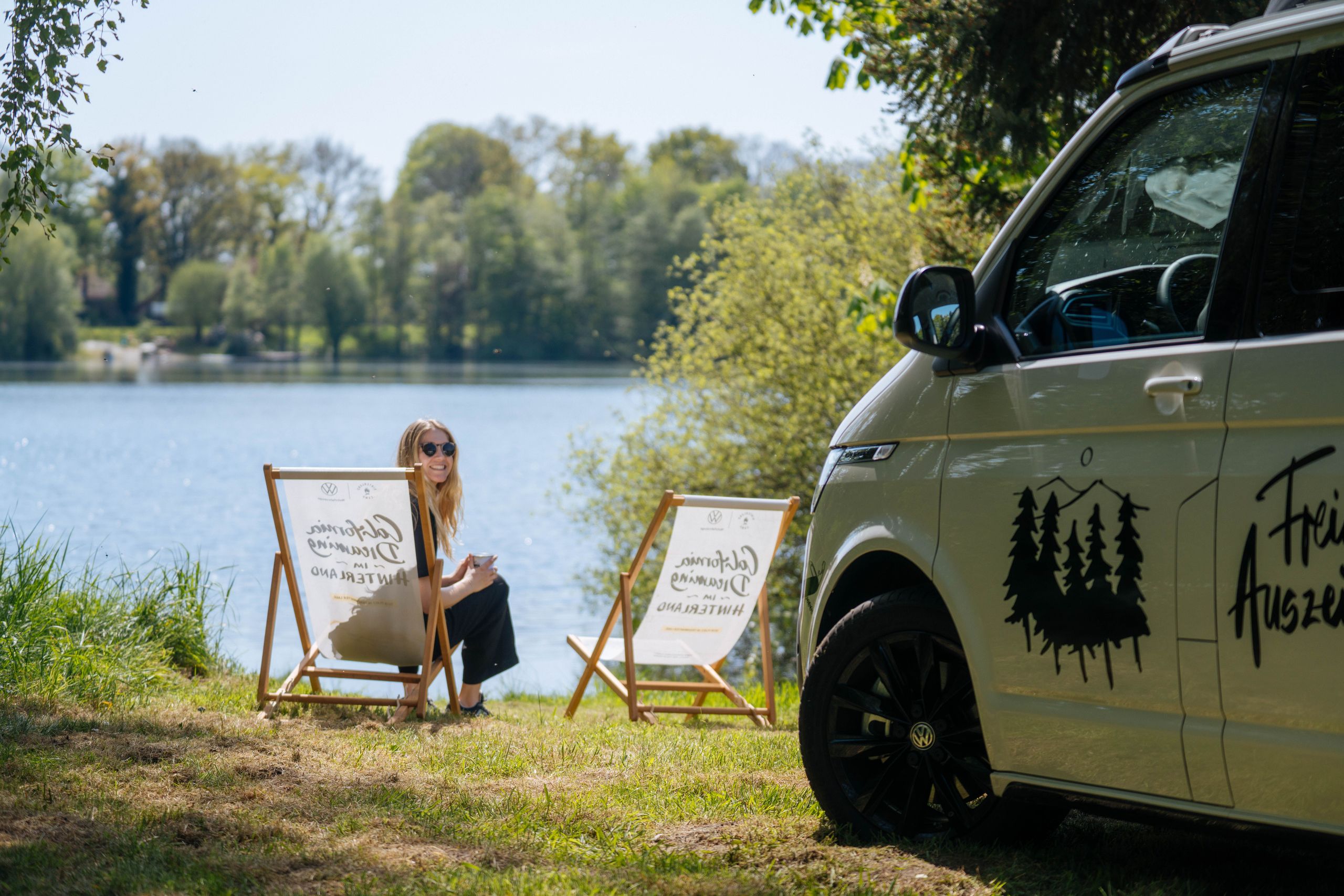 Eine Frau sitzt in einem Stuhl direkt am See vor ihrem Camper, der auf einem Camping Stellplatz direkt am See steht