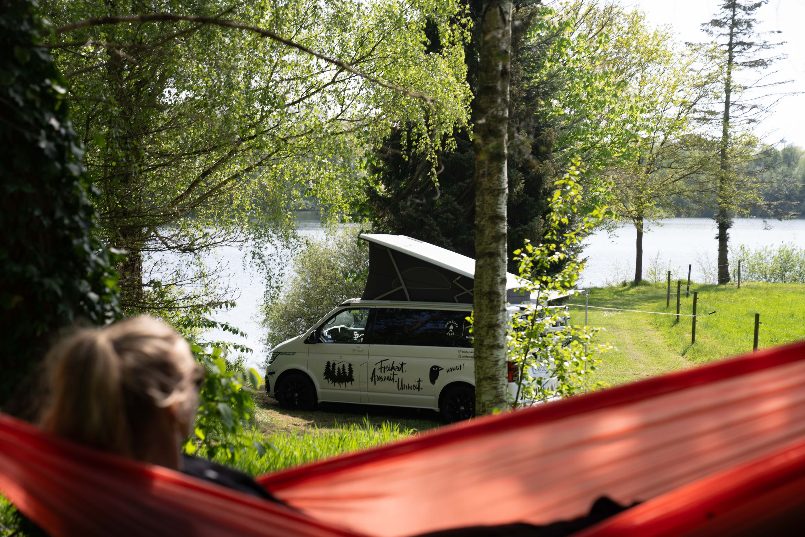 Frau liegt in einer Hängematte vor einem VW-Camper, der auf einem Wohnmobilstellplatz direkt am See steht