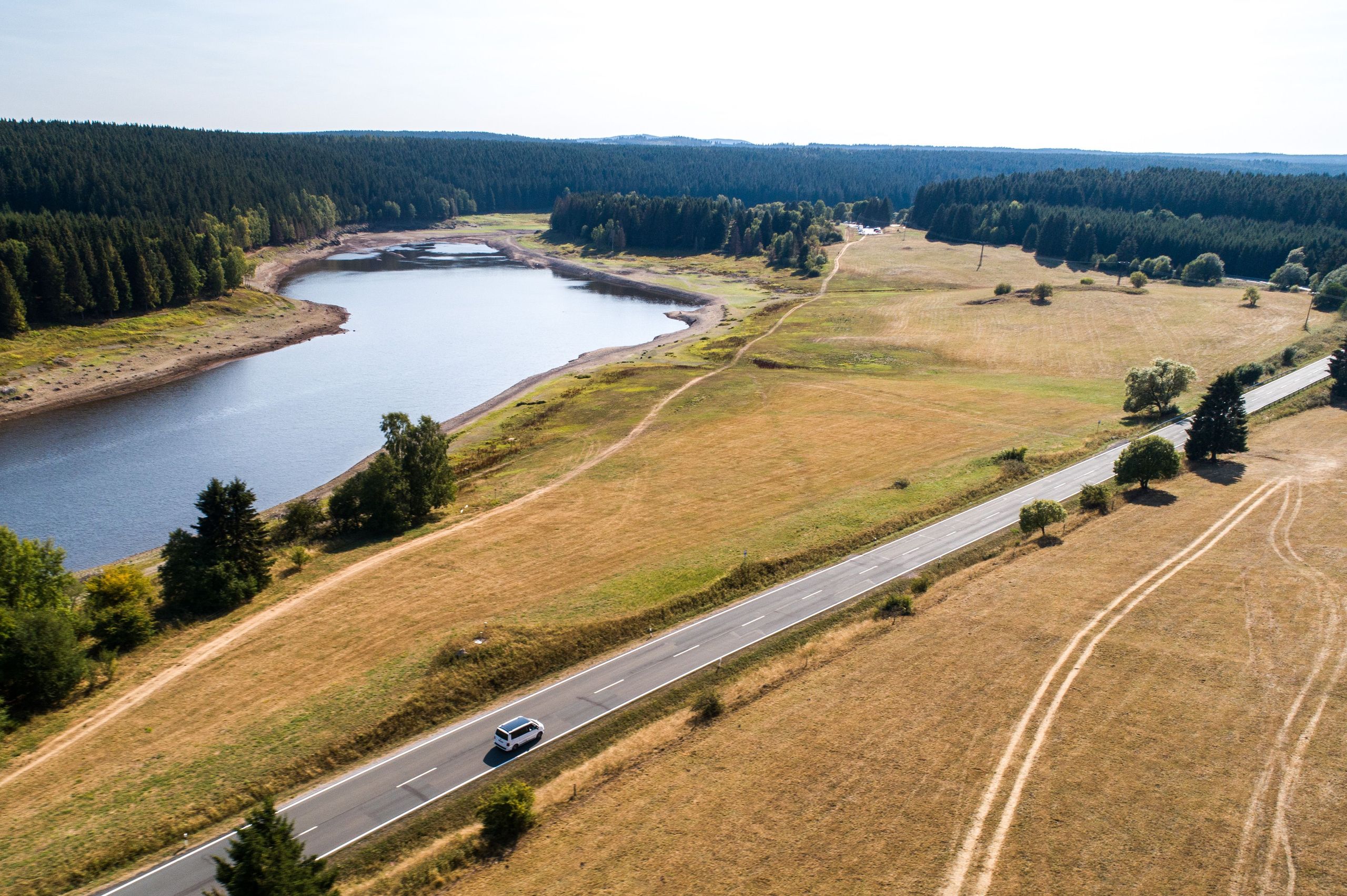 Eine Drohnen-Aufnahme vom Harz. Man erkennt ein Auto das auf dem Weg zu einem Zeltplatz von hinterland.camp ist, welches an einem großen Fluss vorbei und entlang eines Waldes fährt.