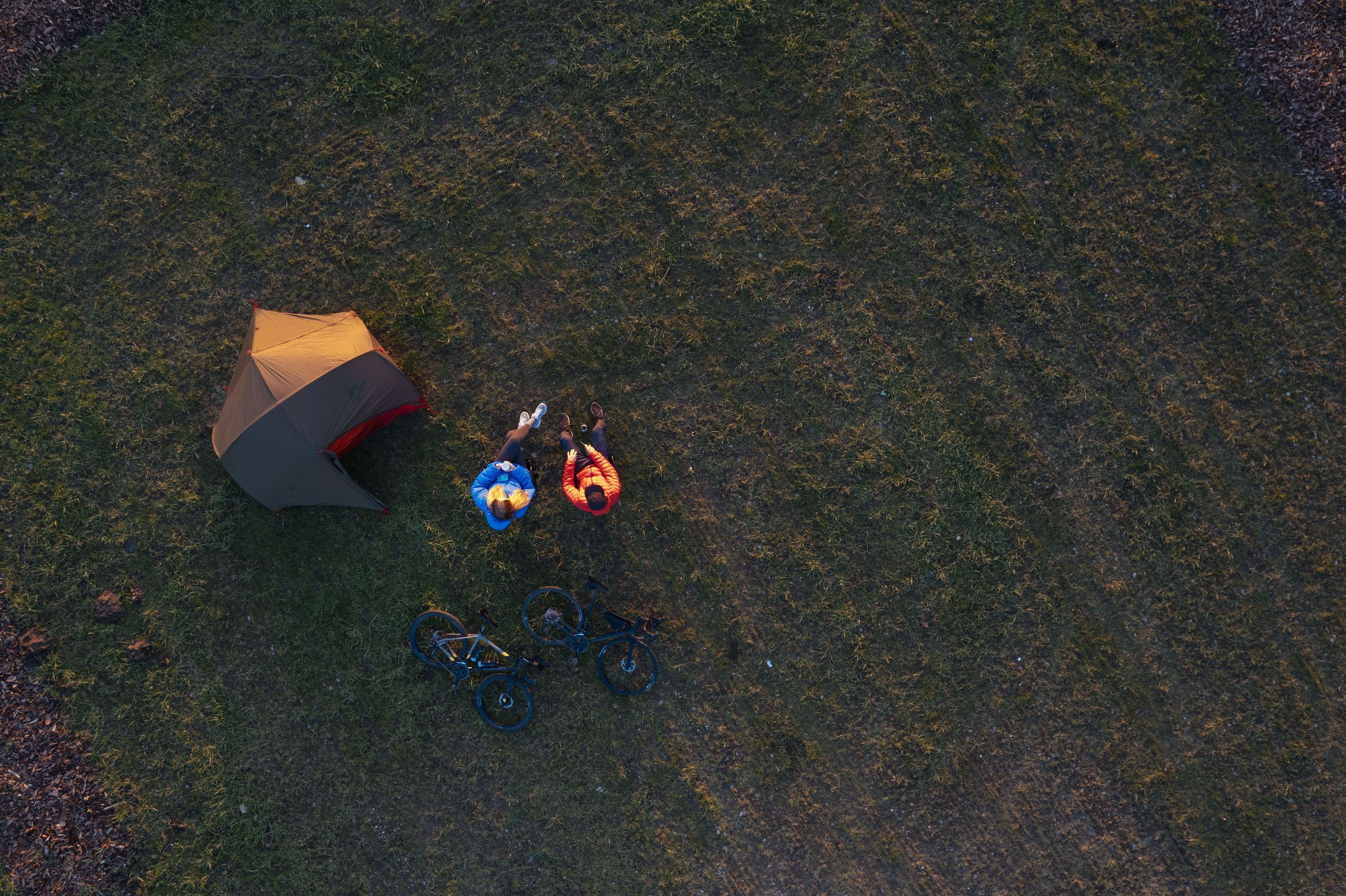 Vogelperspektive auf eine Frau und einen Mann, die im Campingstühlen neben ihrem Zelt bei Sonnenuntergang sitzen. Sie zelten auf einem naturnahen Zeltplatz in der Nähe von Hamburg.