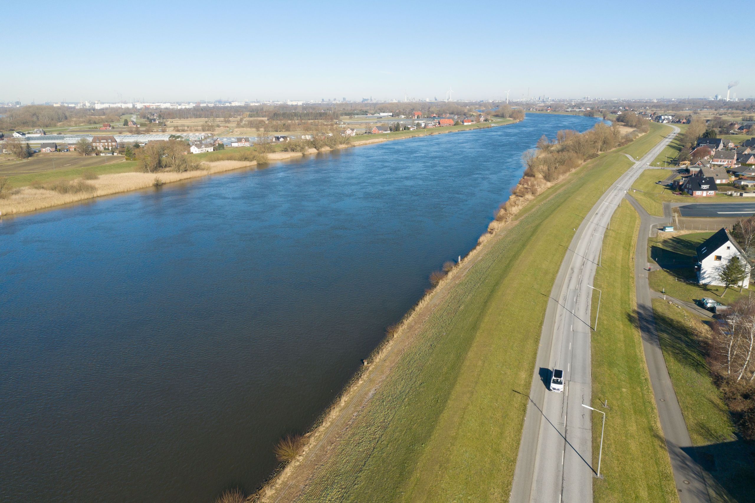 Drohnen-Aufnahme Camper fährt auf einer Straße entlang der Elbe, kleines Wohngebiet und Wiesen