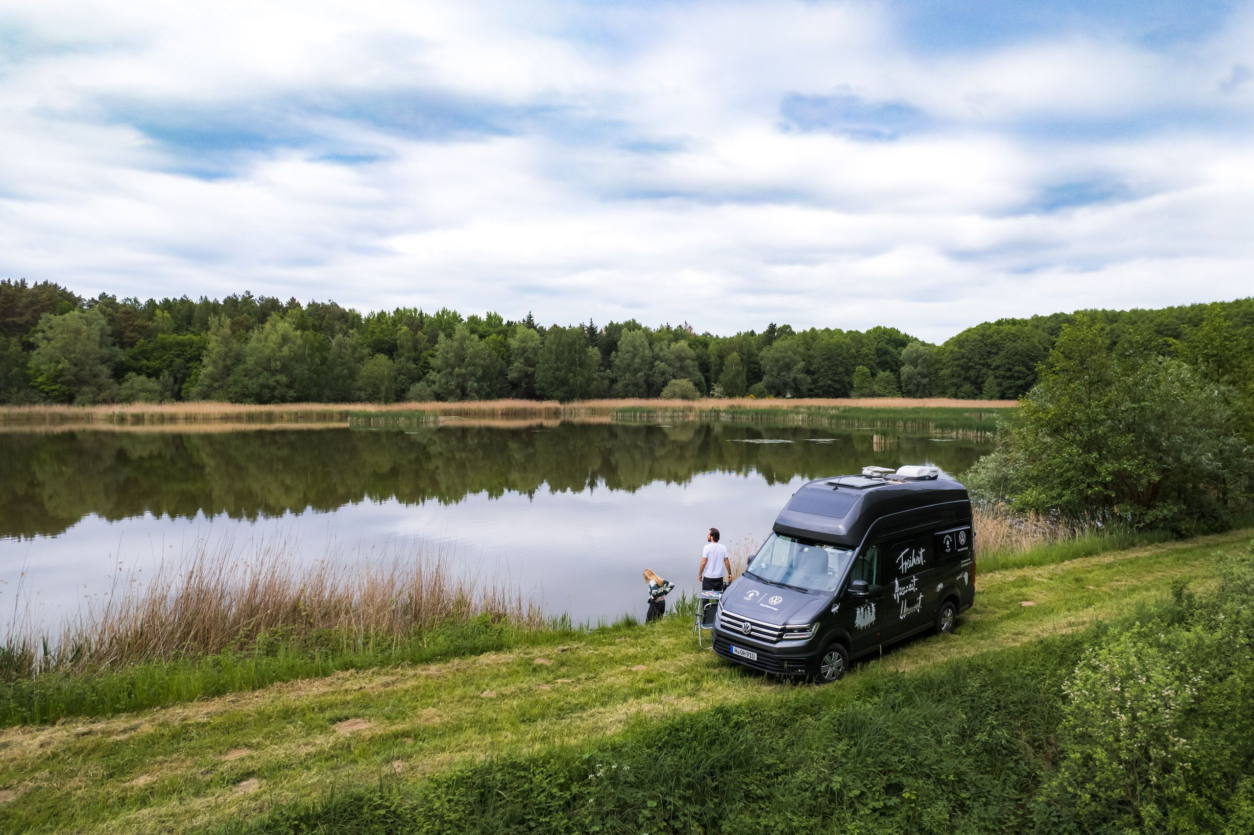 VW-Camper steht auf einem Naturcampingplatz von hinterland.camp direkt am See, umgeben von Wald und Wiesen