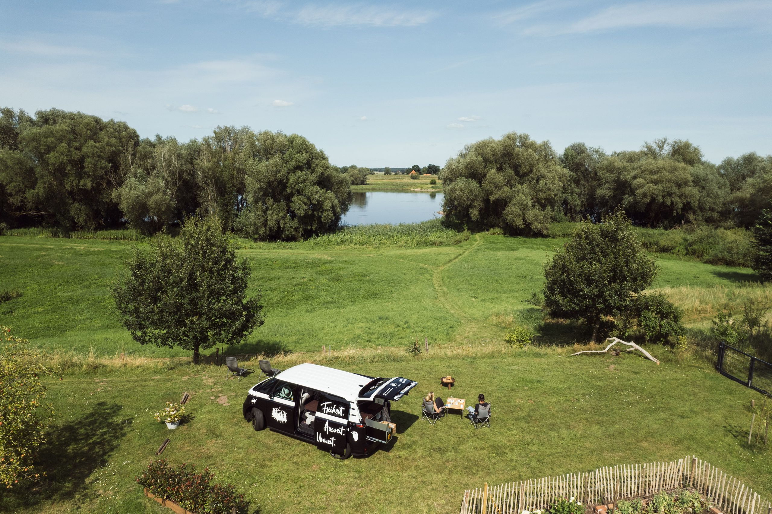 Ein ID-Buzz mit Campingbox steht auf einem Minicamping in Alleinlage direkt an der Wasserkante der Elbe. Vor dem Van sitzen eine Frau und ein Mann am Campingtisch. Den Mini-Campingplatz kann man auf hinterland.camp buchen.