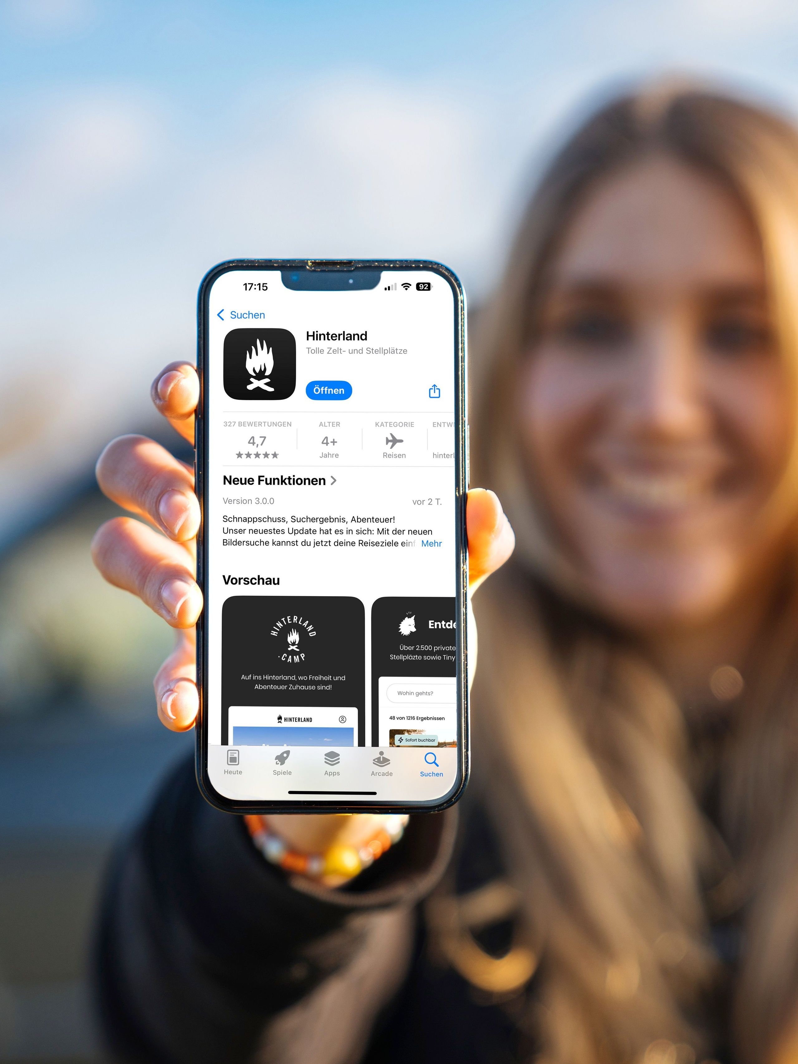Die Kamera schaut über die Schulter einer Frau, die ein Hand in der Hand hält, auf dem die Hinterland-Camping-App geöffnet ist.