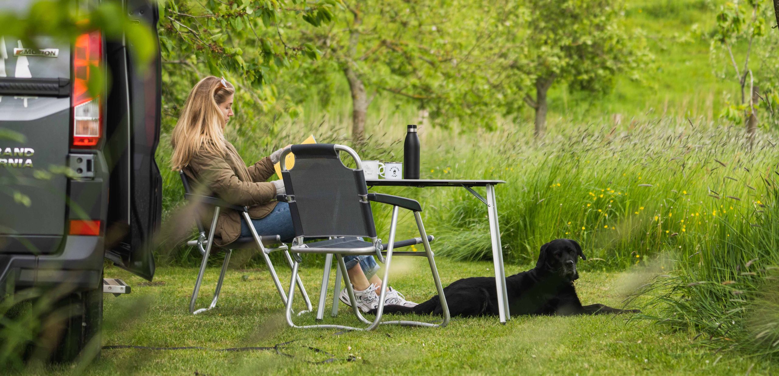 Camper steht auf einem eingezäunten Stellplatz im hohen Gras umgeben von Bäumen, man erkennt den Hund und seine Besitzerin vor dem Camper sitzen