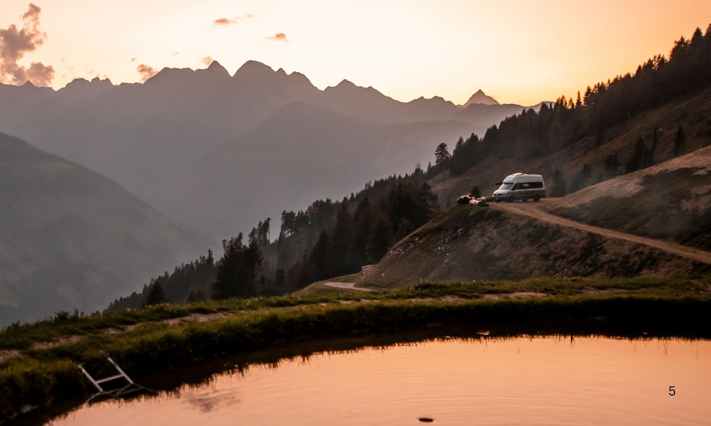 Ein Camper steht auf einem Berggipfel inmitten der Alpen Österreichs. Im Vordergrund spiegelt sich der Sonnenuntergang auf der Oberfläche eines kleinen Sees.