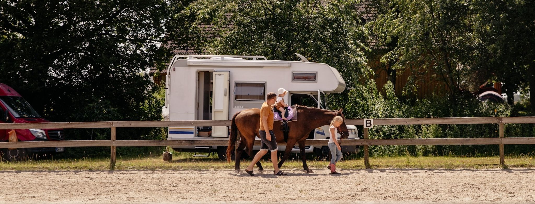 Ein Camper steht im Hintergrund und vorne reitet ein kleines Kind auf einem Pferd auf einem Abenteuer-Campingplatz von hinterland.camp