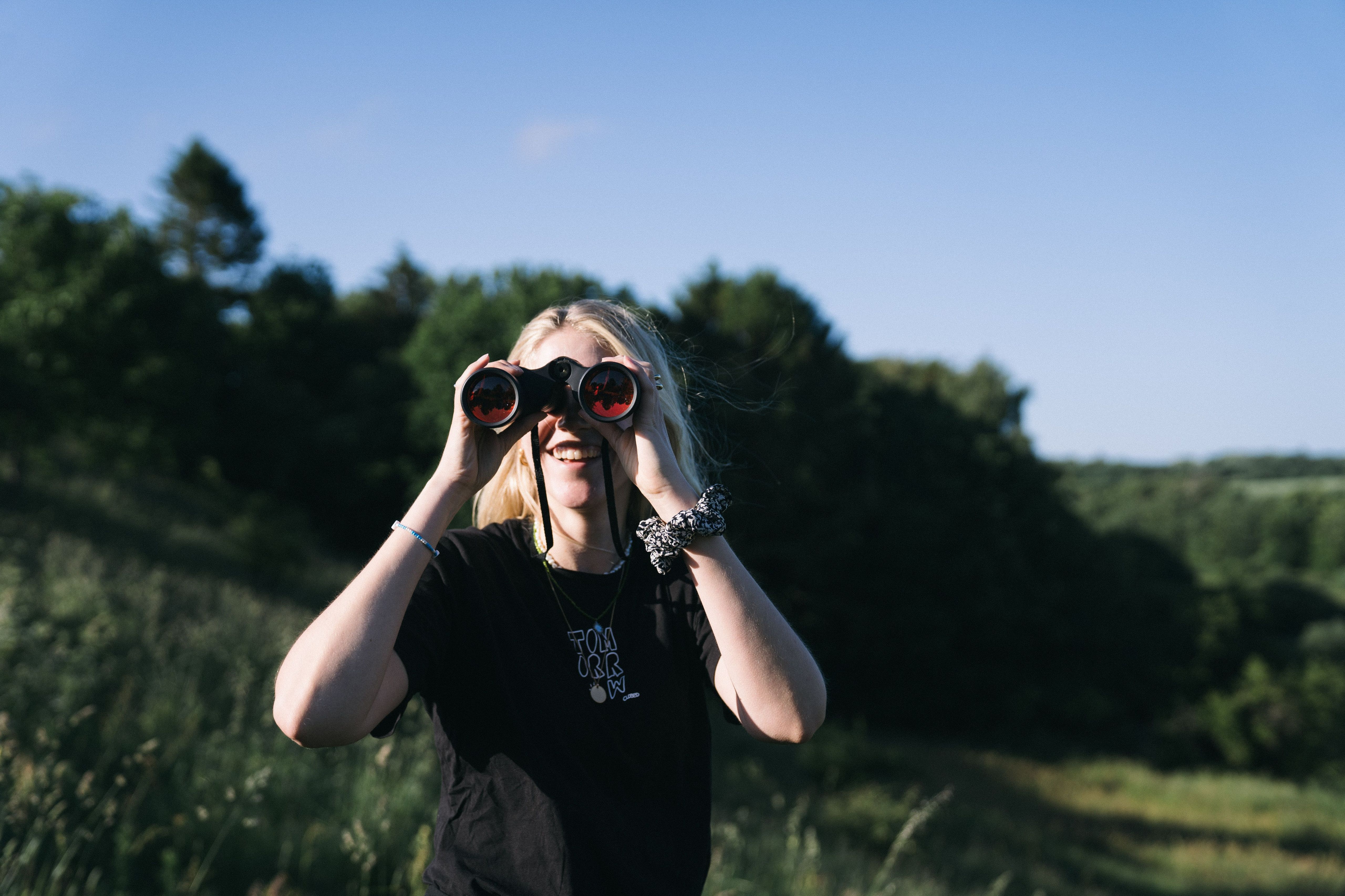 Frau steht mit einem Fernglas an einem Tal. Sie befindet sich vor einer kleinen Hütte im Wald, die man per hinterland.camp buchen kann.