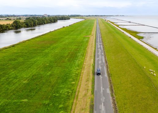 T3 Camper fährt über eine Straße in Niedersachsen, die von Wasser, grünen Wiesen, einem Fluss und Bäumen umgeben ist