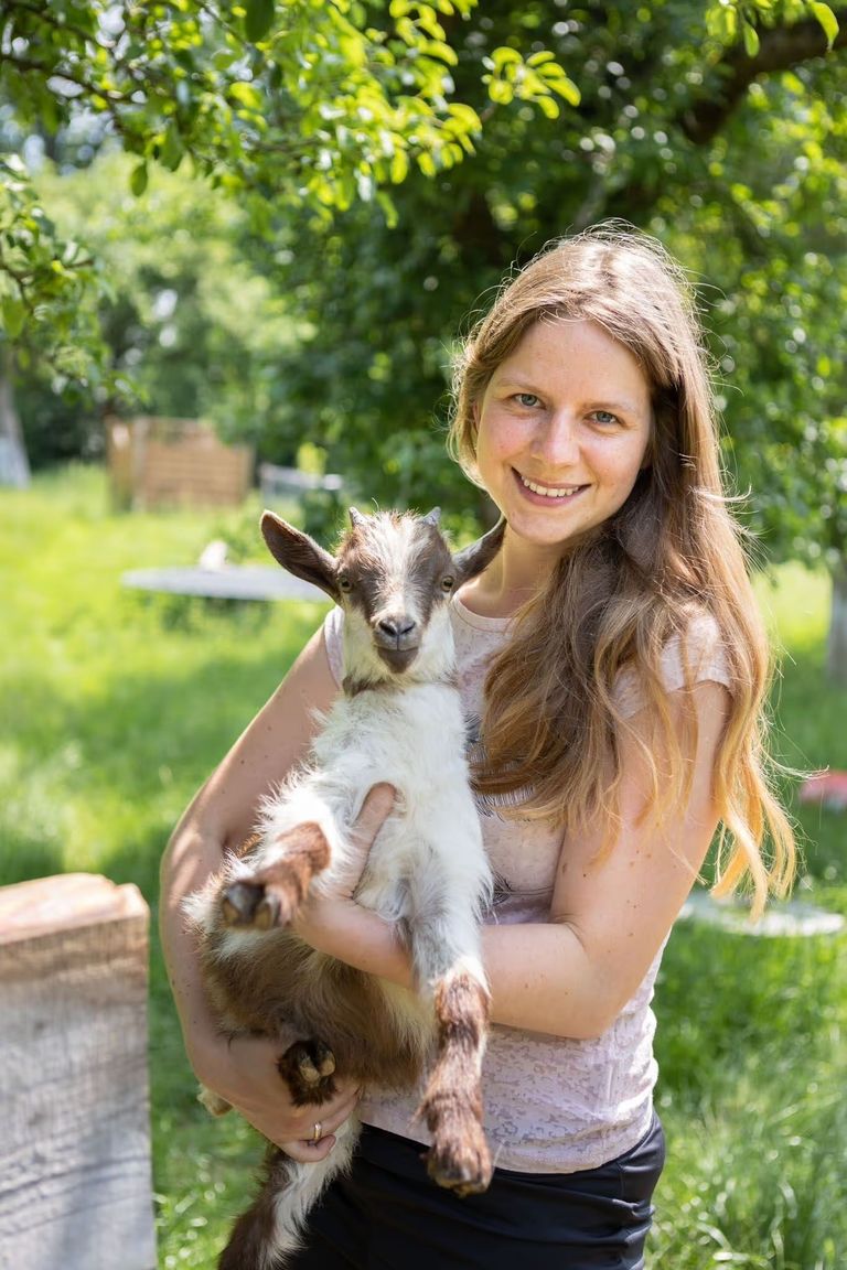 Eine Frau steht auf einer grünen Wies und hat eine Zwergziege auf dem Arm. Die Wiese gehört zu dem Abenteuer-Campingplatz, den man auf hinterland.camp buchen kann.