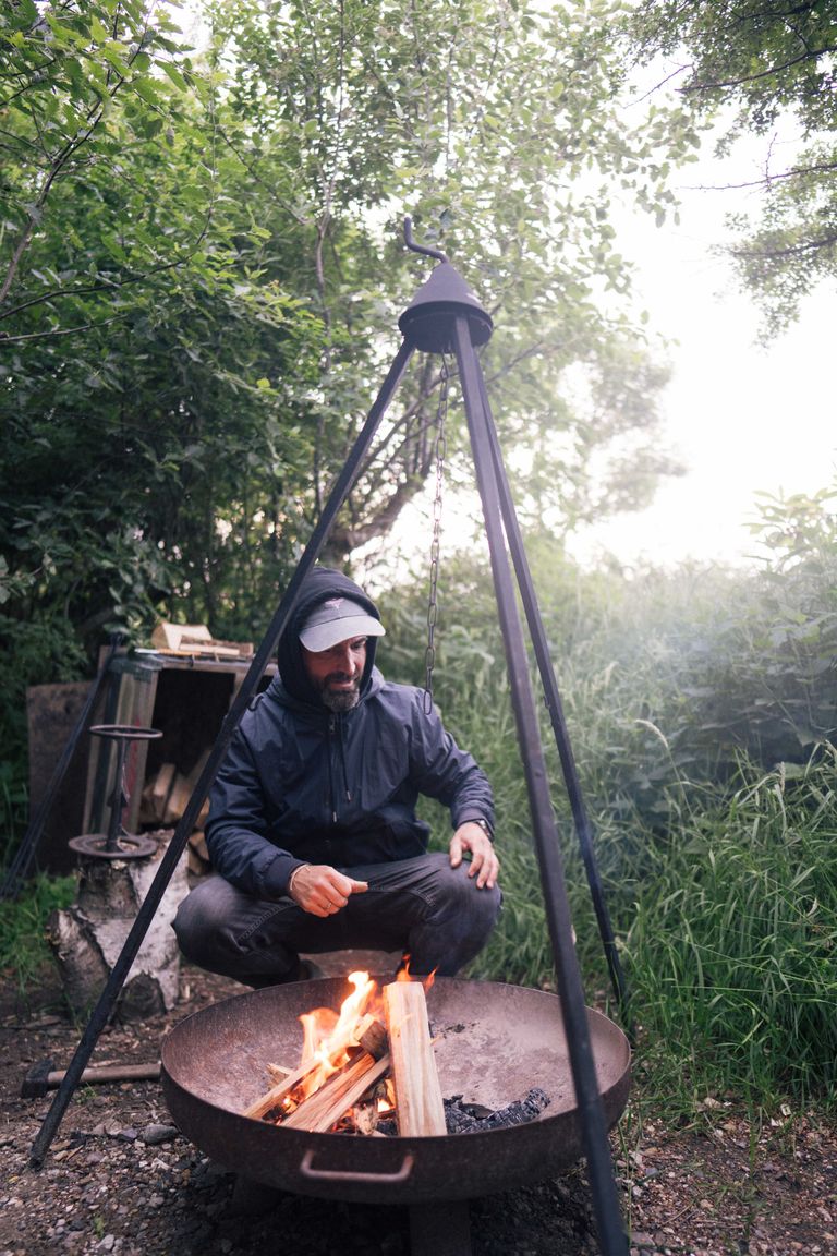 Ein Mann benutzt den Schwenkgrill einer gemütlichen Hütte im Wald mit Panoramablick, die man bei hinterland.camp buchen kann