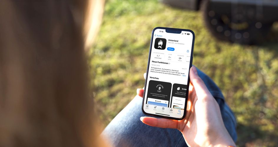 Die Kamera schaut über die Schulter einer Frau, die ein Hand in der Hand hält, auf dem die Hinterland-Camping-App geöffnet ist.
